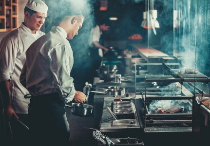 Food concept. Chef in white uniform monitors the degree of roasting and greases meat with oil in saucepan in interior of modern restaurant kitchen. Preparing traditional beef steak on barbecue oven.